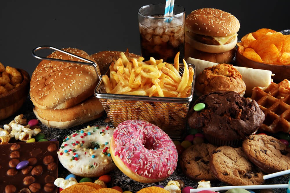 Fotografia de fast food e ultraprocessados: hambúrgueres, batata frita, refrigerante, salgadinhos, donuts e biscoitos, sem marcas comerciais visíveis.